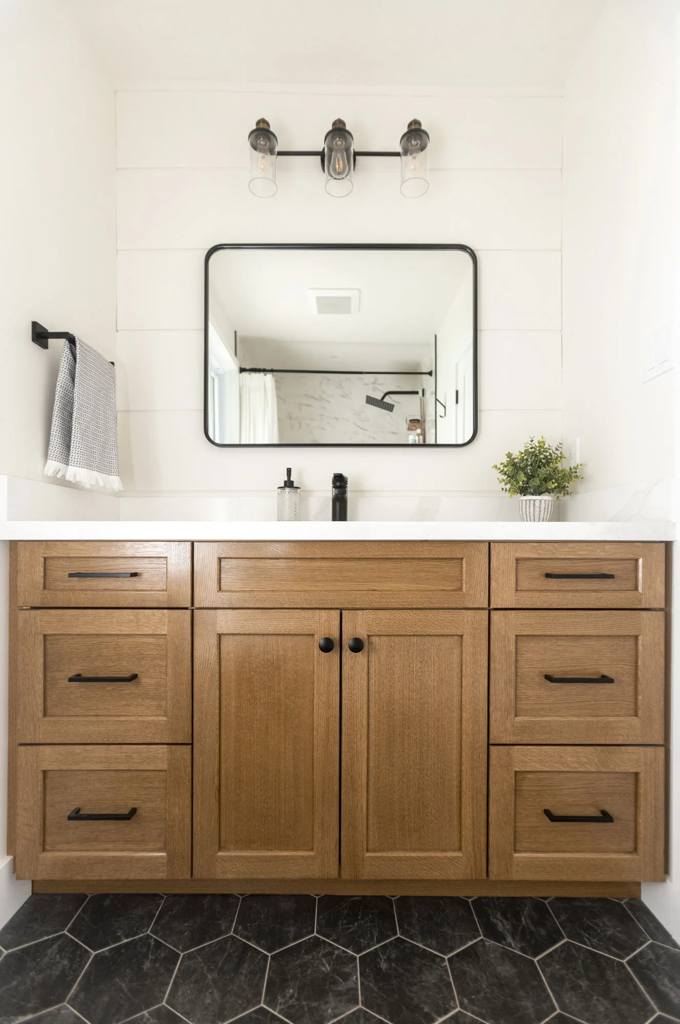 a bathroom cabinet accented with black handles and a black trimmed rectangular mirror with a vintage 3 light overtop