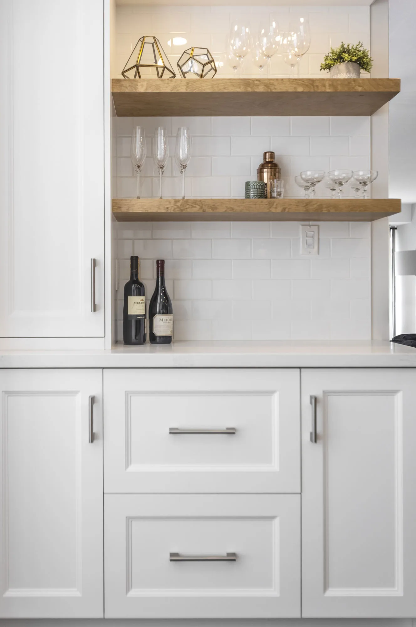 A close up of an area of a kitchen with white cupboards with silver handles and 2 wooden shelves with a white backsplash