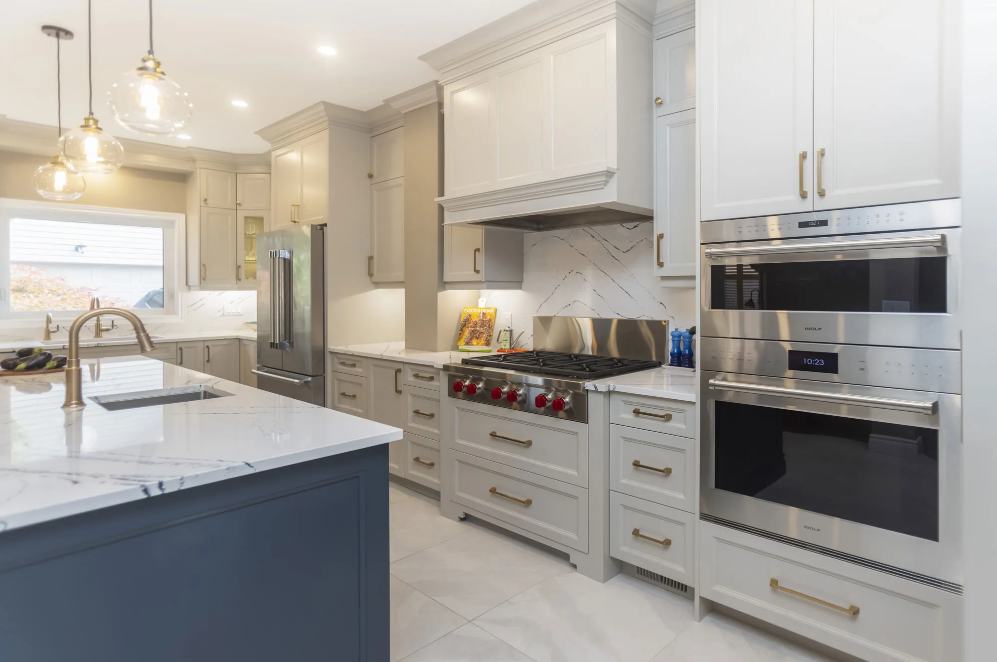 A modern kitchen with off-white cabinentry accented with gold handles along with a dark blue island with white counter tops and gold fixtures and stainless steel applicances