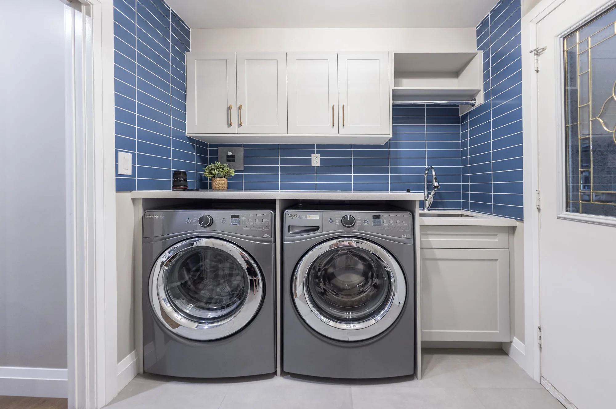 A small laundry room area with stainless steel washer and dryer with a blue subway tile back splash and white cabinets over top