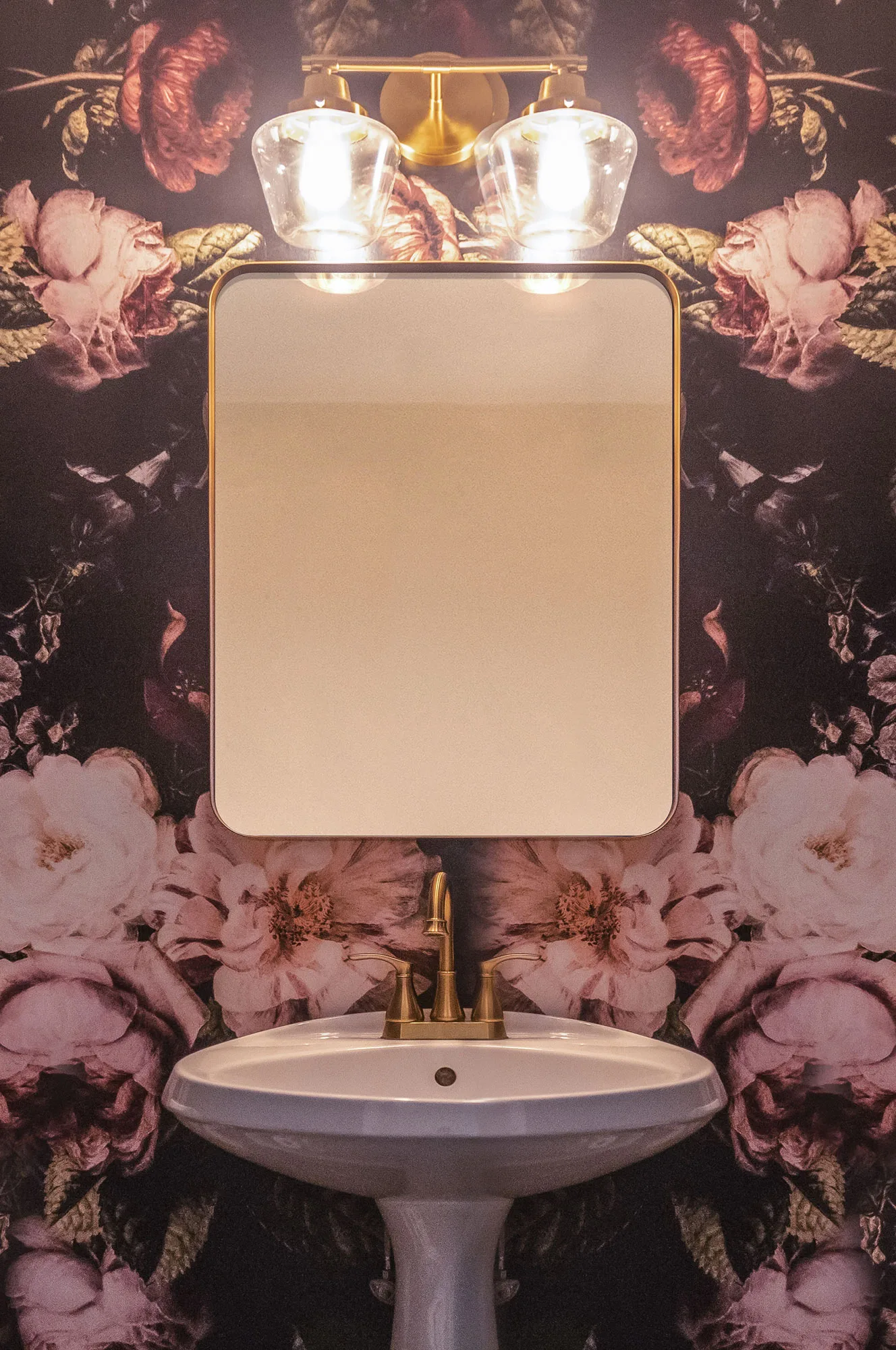 A close up of a pedastal sink and a rounded gold trimmed mirror with large pink floral wallpaper