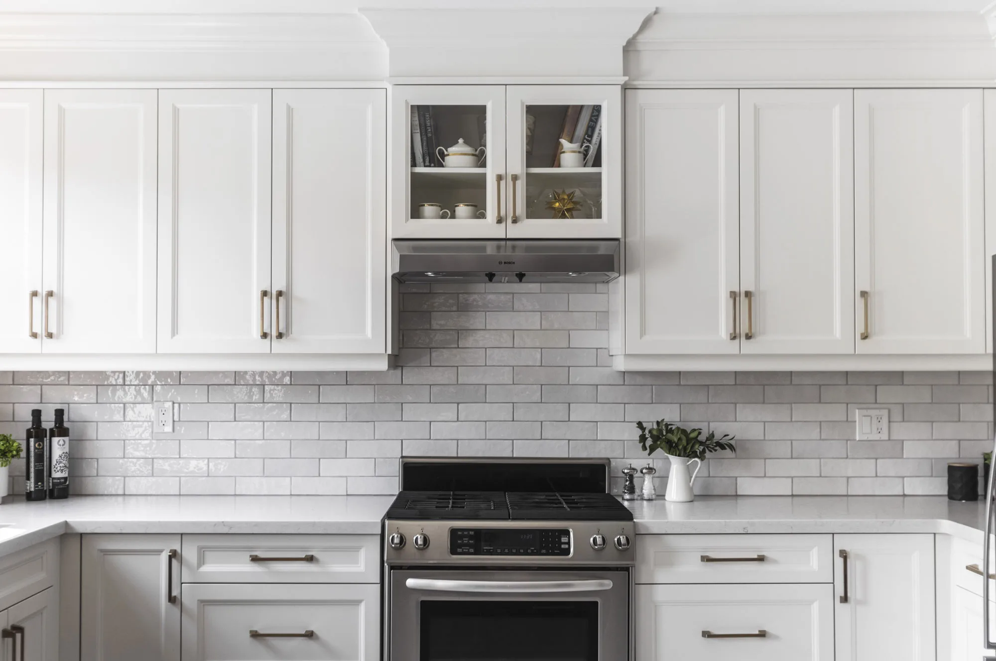 A kitchen with white upper and lower cupboards with silver handles with a stainless steel oven and hood with white and light grey subway tile backsplash