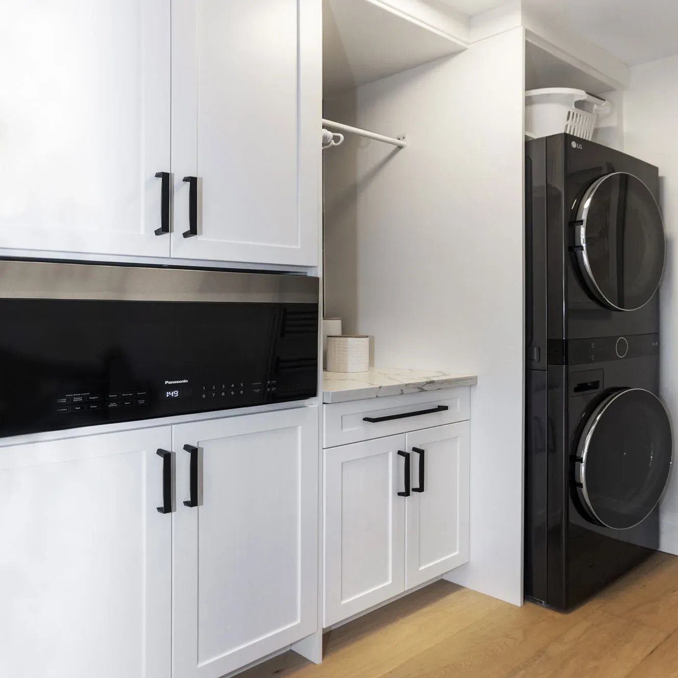 A laundry room with white cabinets trimmed with black handles along with an area to hang clothes and black stackable washer/dryer.