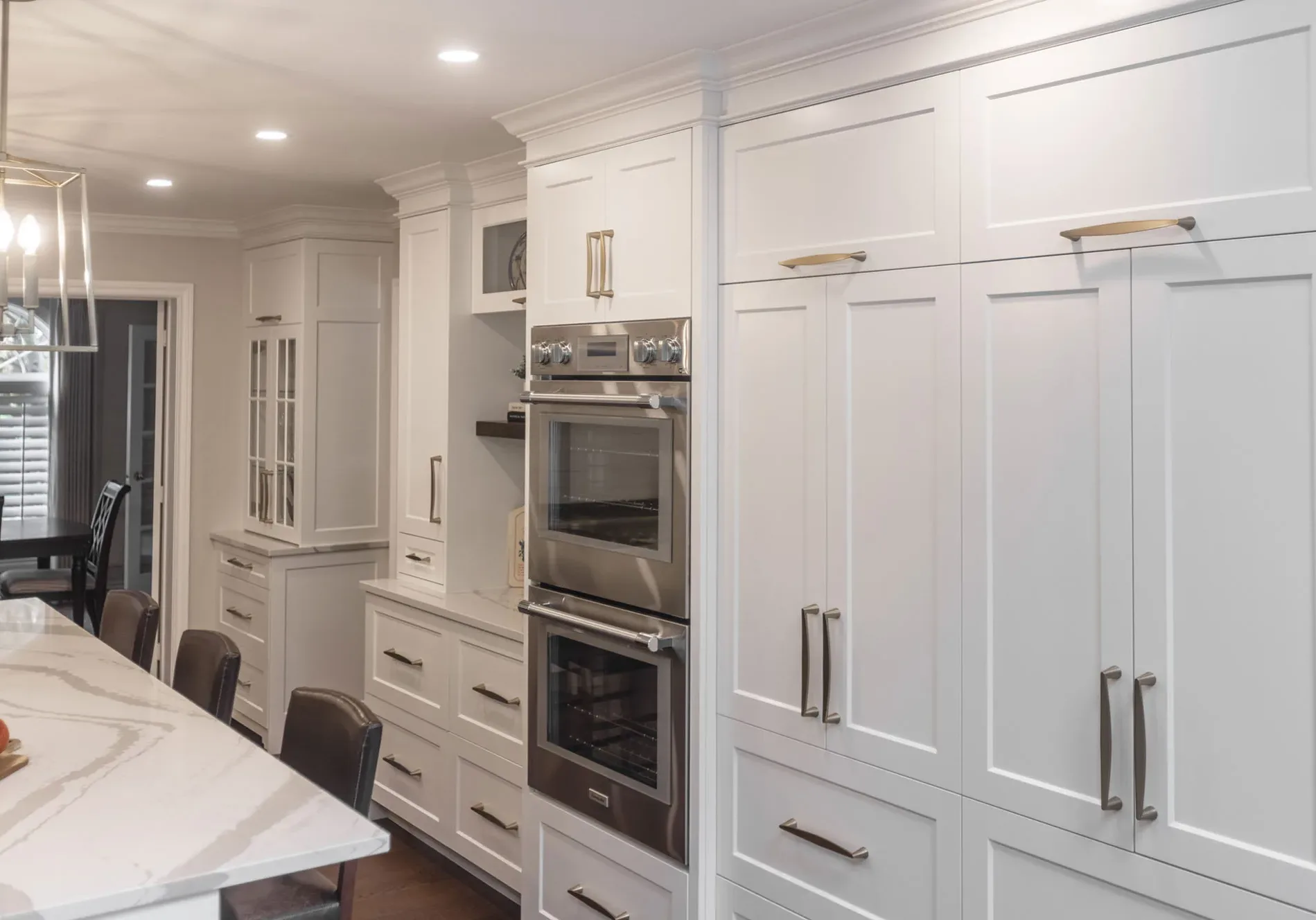 A large section of a wall in a kitchen with white cupboards with large silver handles with stacked ovens