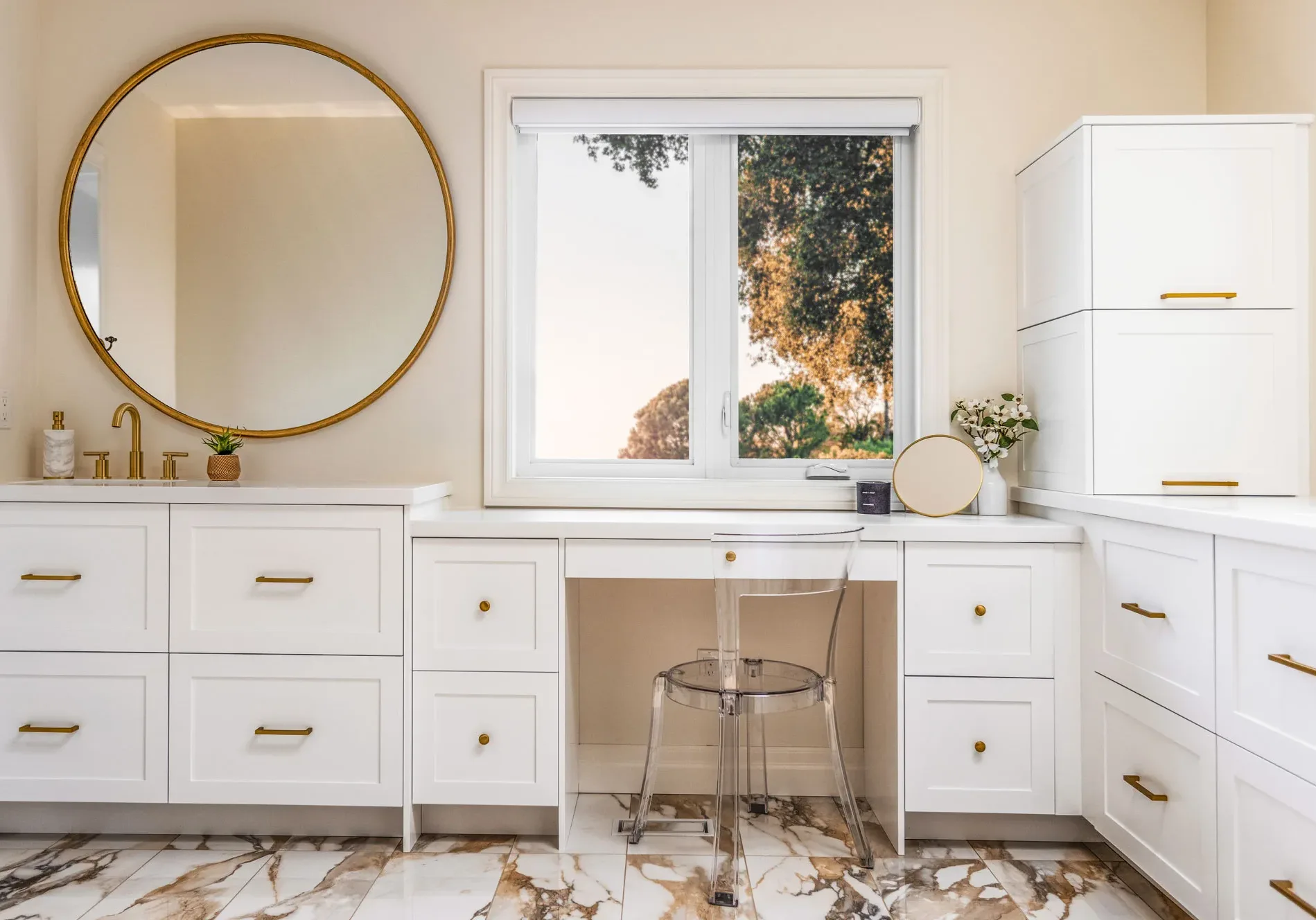 A contemporary bathroom featuring white cabinets with gold handles, a seating area adorned with a transparent plastic chair, and a sink topped with a circular mirror accented with gold trim.